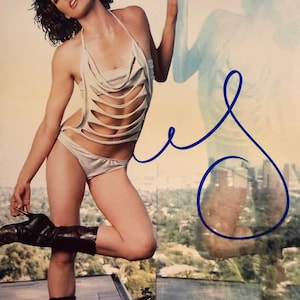 May include: A woman in a silver halter top with cut-out details and matching briefs, and black boots. The image features a blue ink signature and a cityscape background.
