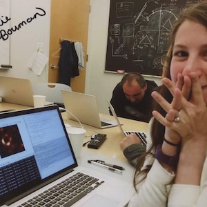 May include: A collaborative workspace with several laptops in use. One screen shows a scientific image. The name "Katie Bouman" is handwritten on a whiteboard. A person is smiling, with hands near their face, looking at the camera.