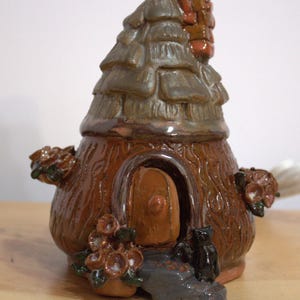 May include: A ceramic fairy house shaped like a tree stump with a brown door, a gray roof, and brown and green floral accents. A black cat figurine sits on the steps.