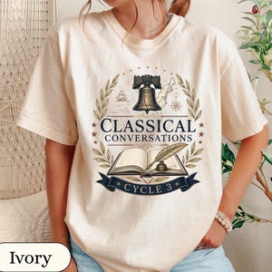 May include: An ivory short-sleeve t-shirt featuring a graphic print of the Liberty Bell, an open book with a quill pen, and the text "CLASSICAL CONVERSATIONS" and "CYCLE 3" framed by laurel branches, stars, a ship, and a compass rose.