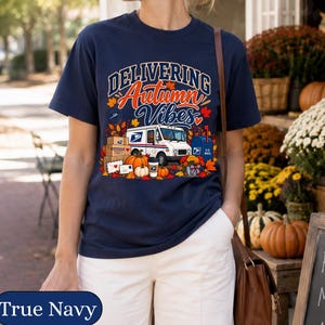 May include: A navy blue t-shirt featuring a graphic print with the text "DELIVERING Autumn Vibes". The design includes a white mail truck, stacked cardboard boxes, a blue mailbox, orange pumpkins, and colorful autumn leaves in shades of red and gold.