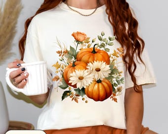 May include: Ivory-coloured t-shirt with a watercolour design of pumpkins, daisies, and foliage. The pumpkins are orange, the daisies are white, and the leaves are green and brown. The word "Ivory" is printed on a label at the bottom.
