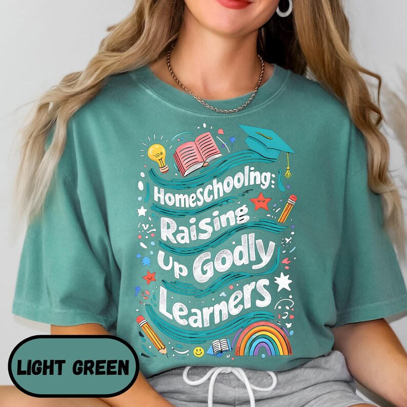May include: Light green t-shirt with the text "Homeschooling: Raising Up Godly Learners" in a whimsical design. The design includes a lightbulb, open book, graduation cap, pencils, stars, and a rainbow. The shirt is a solid light green color.