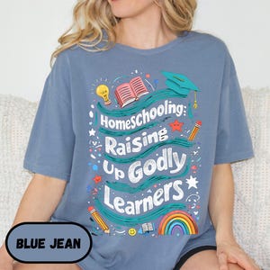 May include: Blue jean colored t-shirt with the text "Homeschooling: Raising Up Godly Learners" in white. The design includes a graduation cap, open book, rainbow, and pencils. The shirt also has the text "BLUE JEAN" in a blue oval.