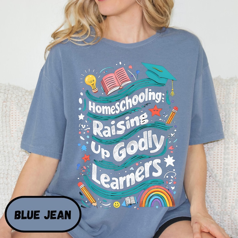 May include: Blue jean colored t-shirt with the text "Homeschooling: Raising Up Godly Learners" in white. The design includes a graduation cap, open book, rainbow, and pencils. The shirt also has the text "BLUE JEAN" in a blue oval.