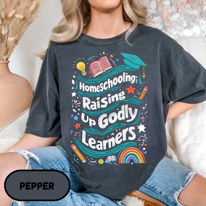 May include: Dark gray t-shirt with the text "Homeschooling: Raising Up Godly Learners" in a colorful design. The design includes a graduation cap, open book, rainbow, and pencils. The shirt is a comfortable, casual style.