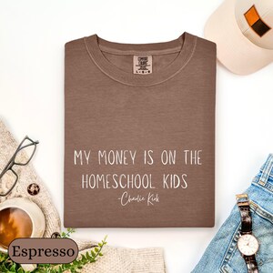 May include: Espresso-colored sweatshirt with the text "MY MONEY IS ON THE HOMESCHOOL KIDS - Charlie Kirk" in white. The sweatshirt is folded and surrounded by accessories, including a watch, glasses, and a hat.
