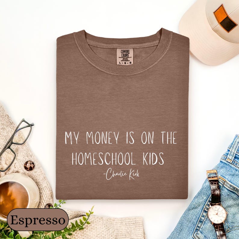 May include: Espresso-colored sweatshirt with the text "MY MONEY IS ON THE HOMESCHOOL KIDS - Charlie Kirk" in white. The sweatshirt is folded and surrounded by accessories, including a watch, glasses, and a hat.