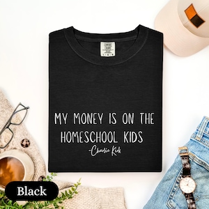 May include: Black crew neck t-shirt with the text "MY MONEY IS ON THE HOMESCHOOL KIDS - Charlie Kirk" in white. A white baseball cap, glasses, and a watch are visible in the background.