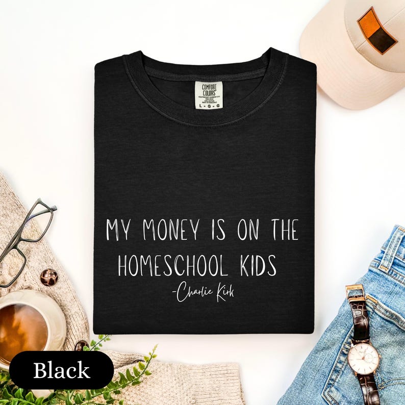 May include: Black crew neck t-shirt with the text "MY MONEY IS ON THE HOMESCHOOL KIDS - Charlie Kirk" in white. A white baseball cap, glasses, and a watch are visible in the background.