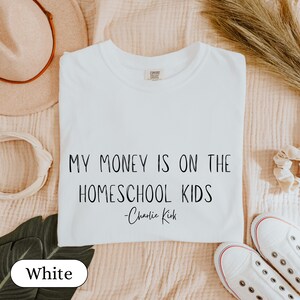 May include: White t-shirt with the text "MY MONEY IS ON THE HOMESCHOOL KIDS - Charlie Kirk" in black. The shirt is displayed on a neutral-toned surface, with white sneakers visible in the bottom right corner.