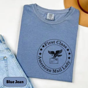Può includere: Una maglietta blu con una grafica nera di un'aquila che trasporta una lettera con il testo "First Class American Mail Lady" in un cerchio con stelle.