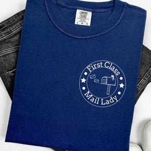 Puede incluir: Camiseta azul marino con un diseño circular blanco que dice "First Class Mail Lady". La camiseta está doblada y colocada sobre unos vaqueros oscuros. La camiseta tiene una etiqueta Comfort Colors.