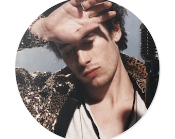 May include: A circular image of a person with their hand on their forehead, wearing a gold sequined jacket and holding a vintage microphone. The person is wearing a white shirt and a gold necklace. The background is a light blue.