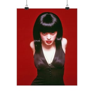 May include: A poster featuring a woman with black hair and red lipstick against a red background. She wears a black dress and a silver necklace. The image is attached to a wall with a black clip.