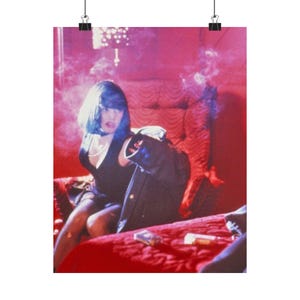 May include: A poster print showing a woman with blue hair in a black dress, seated on a red bed. She holds a black jacket. Cigarettes and smoke are visible, creating a vintage aesthetic.