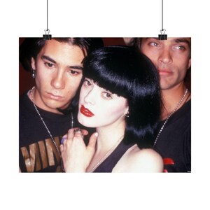 May include: A poster showing three people, two men and a woman, in a close-up. The woman has dark hair and red lipstick. The men have dark hair and are wearing black tops. The poster is held by two black binder clips.