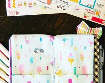 May include: A white notebook with a zipper closure and a colorful pattern of pink flamingos, ice cream cones, popsicles, palm trees, and bicycles. The notebook is open to reveal a blank page and a second page with the same pattern. The notebook is tied together with a white string.