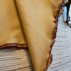 May include: A pair of folded tan linen napkins with a brown scalloped edge. The napkins are on a white and brown woven surface.