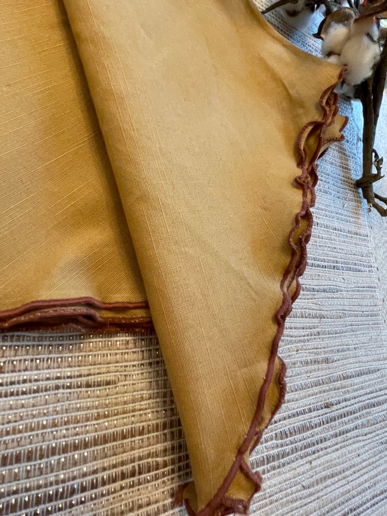 May include: A pair of folded tan linen napkins with a brown scalloped edge. The napkins are on a white and brown woven surface.