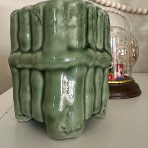 May include: A light green ceramic planter with a textured, vertical ribbed design. The square-shaped planter has a glossy finish and sits on four small feet. A decorative item is visible in the background.