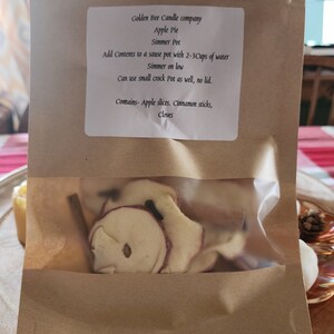 May include: A brown paper bag with a clear plastic window, containing apple slices, cinnamon sticks, and cloves. The bag has a label that reads "Golden Bee Candle company Apple Pie Simmer Pot Add Contents to a sause pot with 2-3Cups of water Simmer on low Can use small crock Pot as well, no lid. Contains- Apple slices. Cinnamon sticks, Cloves"