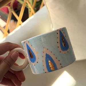 May include: A ceramic mug with a light blue background, adorned with blue and gold teardrop-shaped designs. The mug has a white handle and a slightly angled rim. The design is repeated around the mug's exterior.