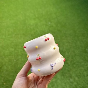 Puede incluir: Maceta de cerámica blanca con un diseño ondulado y un patrón disperso de cerezas rojas y amarillas. La maceta se sostiene en una mano.