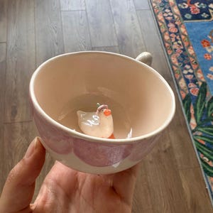 May include: A ceramic mug with a light pink interior and a white and pink exterior design. Inside the mug is a small, white duck figurine with an orange beak and a green hat. The mug is held in a hand.
