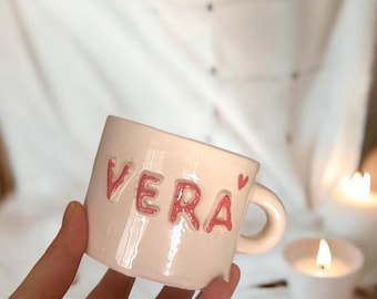 May include: A white ceramic mug with a pink heart and the name "VERA" written in pink letters.