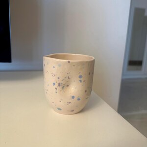 Könnte beinhalten: Eine kleine, hellrosa Keramiktasse mit einem gesprenkelten Design in Blautönen, Lila und Weiß.