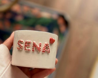 May include: White ceramic mug with the name "SENA" spelled out in red and white candy cane letters. A small red heart is on the right side of the mug.
