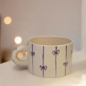 Puede incluir: Una taza de cerámica blanca con un patrón de lazos morados. La taza tiene un asa redondeada y un diseño a rayas.