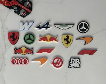May include: A collection of colorful Formula 1 team logos and emblems, including Ferrari, Mercedes, and Red Bull. A toy race car is visible in the upper left corner. The logos are in various shapes and colors, perfect for fans of motorsports.