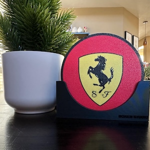 Könnte beinhalten: Ein runder roter Untersetzer mit einem schwarzen und gelben Ferrari-Logo. Der Untersetzer befindet sich in einem schwarzen Halter mit dem Aufdruck "Modern Studio" auf der Unterseite.