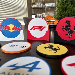 Könnte beinhalten: Eine Sammlung runder Untersetzer mit verschiedenen Logos von Formel-1-Rennteams. Die Untersetzer sind aus einem dicken, strapazierfähigen Material gefertigt und in leuchtenden Farben gehalten. Zu den Logos gehören Red Bull, Ferrari, McLaren, Alpine und Formel 1.