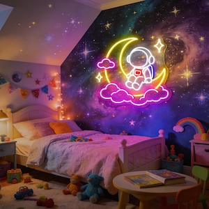 May include: A neon wall sign featuring a white astronaut sitting on a yellow crescent moon surrounded by pink clouds and a four-pointed star. The sign emits a bright glow against a dark wall decorated with a colorful galaxy and star pattern.