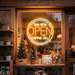 May include: A circular yellow neon sign in a window displays the text "we are OPEN come in" inside a glowing ring. The display includes two small evergreen trees in pots, a gift box with a white ribbon, and a floral arrangement on a wooden ledge.