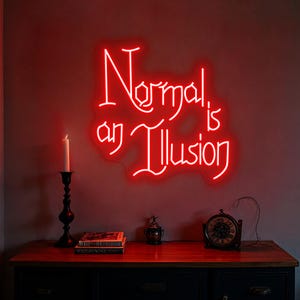 May include: A red neon sign on a wall reads "Normal is an Illusion." Below the sign, a candle in a black candlestick, books, a small decorative container, and a clock sit on a dark wooden surface. The wall is painted a dark red.