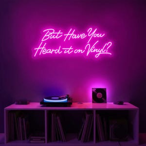 May include: A pink neon sign with the text "But Have You Heard it on Vinyl?" hanging on a purple wall above a white shelf with a record player and vinyl records.
