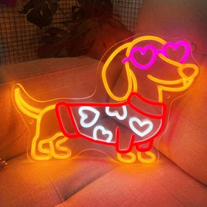 K&ouml;nnte beinhalten: Ein Neon-Schild in Form eines Dackels, der einen roten Pullover mit wei&szlig;en Herzen und rosafarbenen herzf&ouml;rmigen Sonnenbrillen tr&auml;gt. Das Neon-Schild leuchtet in Gelb, Rot und Pink.