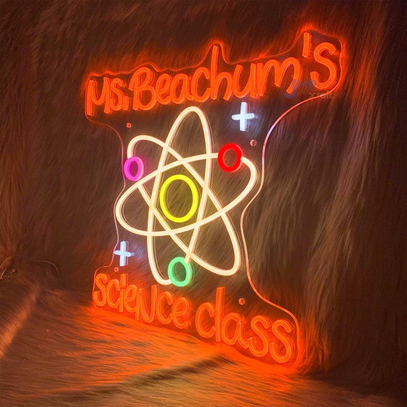May include: Neon sign in the shape of a science class emblem. The sign is orange with the words "Ms. Beachum's science class" in a cursive font. An atom symbol is in the center, with colorful accents.