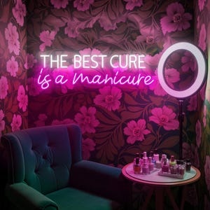 Könnte beinhalten: Ein Maniküre-Salon mit einem Neon-Schild mit der Aufschrift "THE BEST CURE is a manicure". Der Raum hat eine florale Tapete, einen teal-farbenen Samtsessel und einen kleinen Tisch mit Nagellackflaschen. Ein Ringlicht ist rechts positioniert.