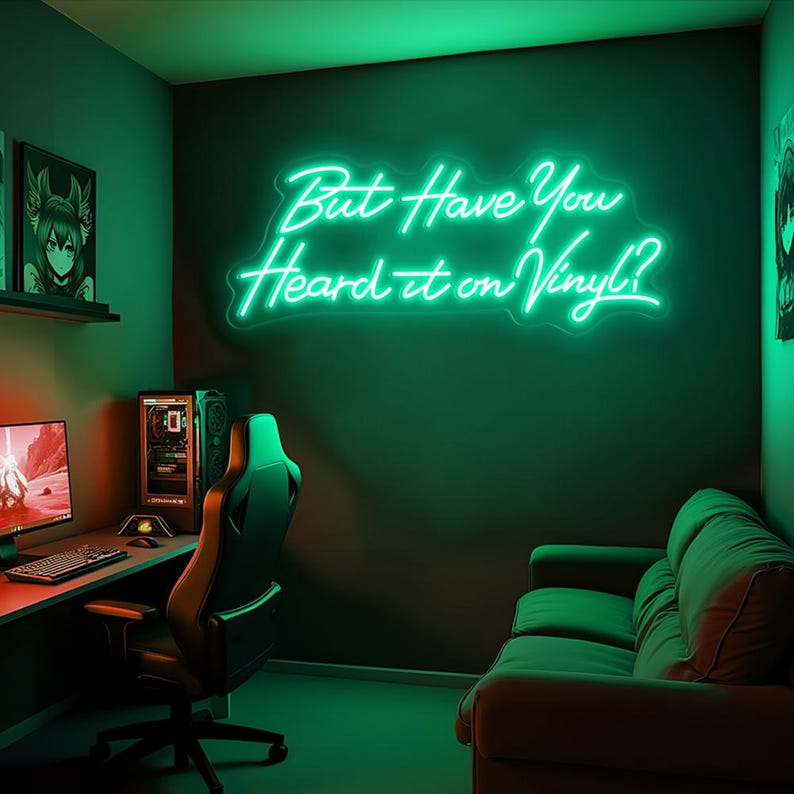 May include: A green neon sign with the text "But Have You Heard it on Vinyl?" hanging on a dark green wall in a room with a sofa and a gaming chair.