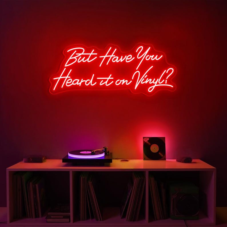 May include: Red neon sign with the text "But Have You Heard it on Vinyl?" hanging on a red wall above a white shelf with a record player and vinyl records.