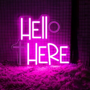 Puede incluir: Un letrero de neón rosa brillante que muestra las palabras "HELLO THERE". El letrero, en una fuente estilizada, está montado sobre un fondo de rejilla oscura. El neón se ilumina con una luz rosa vibrante, creando un efecto visual llamativo. El letrero está colocado sobre una superficie rosa esponjosa.
