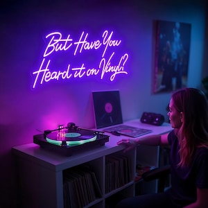 May include: A purple neon sign with the text "But Have You Heard it on Vinyl?" hanging on a wall above a record player. The record player is on a white shelf with a collection of vinyl records.