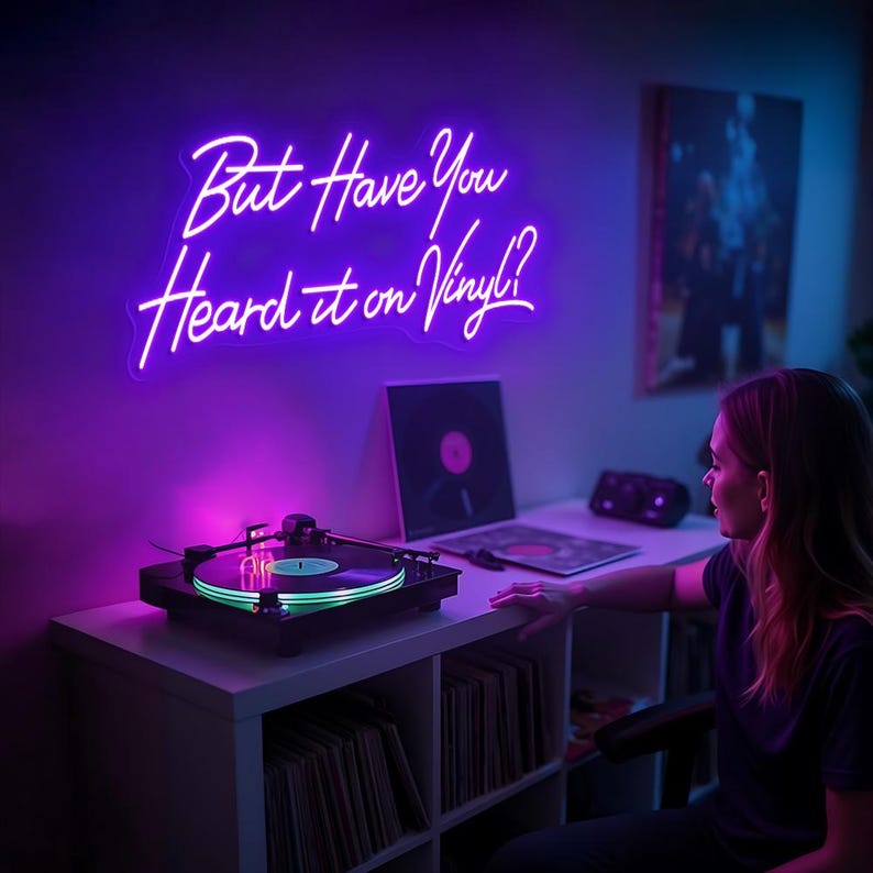 May include: A purple neon sign with the text "But Have You Heard it on Vinyl?" hanging on a wall above a record player. The record player is on a white shelf with a collection of vinyl records.
