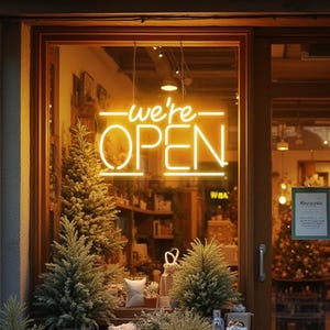 Op de afbeelding: Een etalage toont een heldergeel neonbord met de tekst "we're OPEN". De etalage wordt omlijst door een houten deur en versierd met kleine kerstbomen en andere feestelijke decoraties.