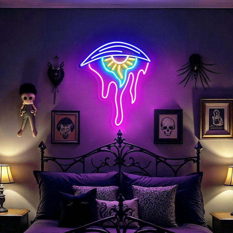 May include: A neon eye-shaped sign with blue, purple, and yellow lights is mounted on a wall above a bed with purple bedding. The room is decorated with framed art and decorative objects, creating a gothic theme.